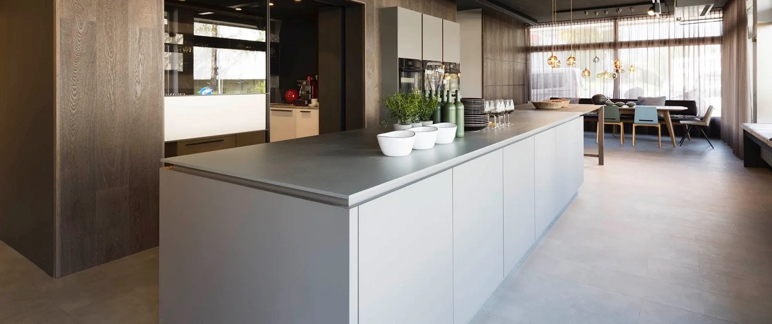 Graue Kochinsel im Showroom, dahinter ein Essbereich vor großen Fenstern